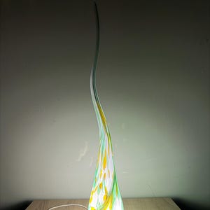 Könnte beinhalten: Eine hohe, schlanke, flammenförmige Glaslampe mit einem wirbelnden Muster in Weiß, leuchtendem Gelb und Hellgrün. Die Lampe hat einen breiten, abgerundeten Sockel, der sich nach oben hin zu einer dünnen, geschwungenen Spitze verjüngt.