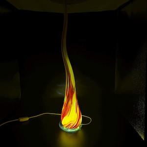 Könnte beinhalten: Eine hohe, schlanke, flammenförmige Glaslampe mit einem wirbelnden Muster aus leuchtendem Gelb und tiefem Rot. Die Lampe steht auf einem runden Sockel und wird von innen beleuchtet, wodurch sie ein warmes Licht ausstrahlt, mit einem weißen Stromkabel.
