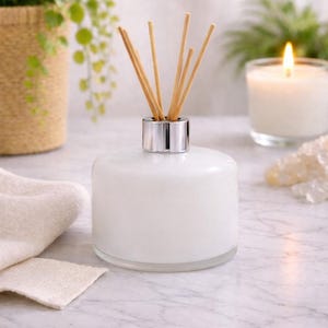 Puede incluir: Un difusor de aceites esenciales de vidrio blanco con cañas de madera y un collar plateado. El difusor se encuentra sobre una superficie de mármol, con una vela encendida y vegetación en el fondo. También hay una pequeña toalla blanca.