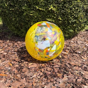 Può includere: Una palla decorativa in vetro giallo con accenti colorati a mosaico. La sfera è posizionata su pacciame marrone, con un cespuglio verde sullo sfondo. Questo ornamento da giardino aggiunge un tocco di colore.