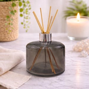 Puede incluir: Un difusor de vidrio gris con un collar plateado, lleno de varillas de madera. La botella está sobre una superficie de mármol, con una vela encendida y vegetación en el fondo. También hay un paño de color neutro.