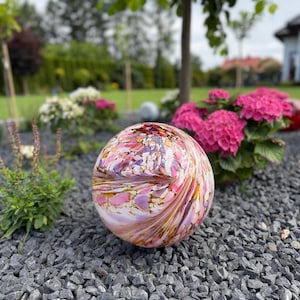 Può includere: Un ornamento sferico in vetro per giardino caratterizzato da un motivo a vortice nei colori rosa, bianco, viola e ambra. La superficie della sfera è liscia e lucida, riflette la luce sulla sua forma arrotondata, creando vivaci sfumature.