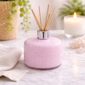 Puede incluir: Un difusor de aceites esenciales de vidrio rosa con un collar plateado y varillas de madera. El difusor está sobre una superficie de mármol, con una vela encendida y vegetación en el fondo. Un artículo decorativo para el hogar.