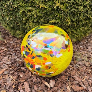 Può includere: Una sfera di vetro gialla con forme irregolari colorate. La sfera decorativa presenta accenti arancioni, blu e verdi. L'ornamento da giardino è posto su un letto di pacciame marrone, con un cespuglio verde sullo sfondo.