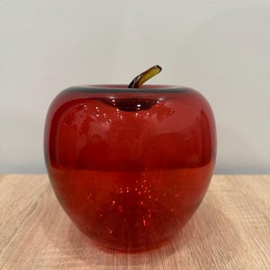 Puede incluir: Una manzana decorativa de cristal rojo con un tallo oscuro. La manzana es de un color rojo vibrante y se encuentra sobre una superficie de madera clara. El cristal tiene un acabado brillante.