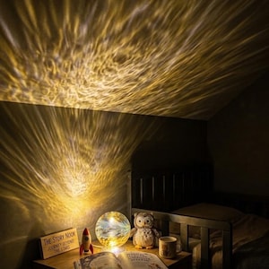 Könnte beinhalten: Eine kugelförmige Lampe wirft ein warmes, wellenförmiges goldenes Lichtmuster an die Wand. Daneben stehen eine kleine Holzrakete, ein Eulen-Plüschtier, eine Tasse und ein offenes Buch. Ein Holzschild trägt die Aufschrift: THE STORY NOOK JOURNEY BEYOND THE STARS.