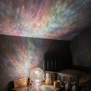 Könnte beinhalten: Eine kugelförmige Mosaikglaslampe, die ein farbenfrohes Licht an die Decke und Wand projiziert. Daneben stehen eine kleine Rakete aus Holz, eine Eulen-Plüschfigur, eine Keramiktasse und ein aufgeschlagenes Buch. Ein Holzschild trägt die Aufschrift THE STORY NOOK JOURNEY BEYOND THE STARS.