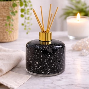 Puede incluir: Un difusor de vidrio negro moteado con un collar dorado que sostiene varias cañas de color claro. La botella se encuentra sobre una superficie de mármol, con una vela encendida y vegetación en el fondo.