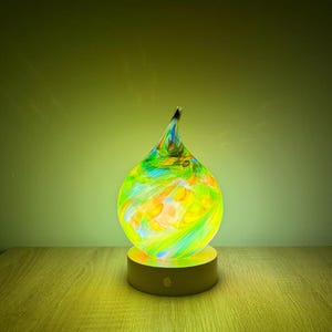 Może przedstawiać: Kolorowa szklana lampa w kształcie łzy, z żywymi odcieniami zieleni, żółci i pomarańczy. Lampa stoi na okrągłej, drewnianej podstawie, oświetlona delikatną poświatą. Lampa ma około 15 cm wysokości.