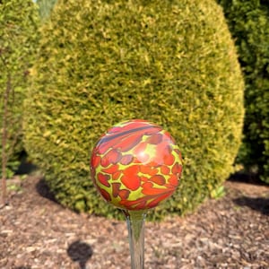May include: A decorative glass garden gazing ball with a clear stem. The ball is a vibrant mix of red, orange, and yellow, with a speckled pattern. The garden ornament is set against a backdrop of green foliage and brown mulch.