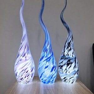 Op de afbeelding: Drie hoge, druppelvormige glazen sculpturen met langgerekte, gebogen toppen. Elk stuk heeft een eigen kleurpatroon: één in lavendel en wit, één in felblauw en wit, en één in zwart en wit, allemaal met een gemarmerd effect op het glas.