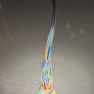May include: A tall, slender, hand-blown glass sculpture lamp with a teardrop-shaped base that tapers into a long, curved neck. The glass features a vibrant, multi-coloured confetti pattern with streaks of blue, red, yellow, and green on a clear base.