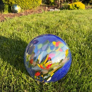 Könnte beinhalten: Dekorative Glaskugel mit Mosaikdesign. Die Kugel hat eine tiefblaue Grundfarbe mit bunten Akzenten in Gelb, Rot und Grün. Sie liegt auf grünem Gras und reflektiert die Umgebung. Durchmesser ca. 20 cm.
