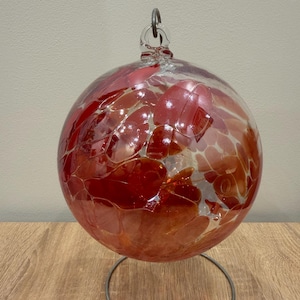 Peut inclure: Une sphère en verre transparent avec des accents rouges et oranges, suspendue par une boucle métallique. La sphère est soutenue par un support métallique circulaire, créant un élément décoratif. Le verre a un aspect texturé.