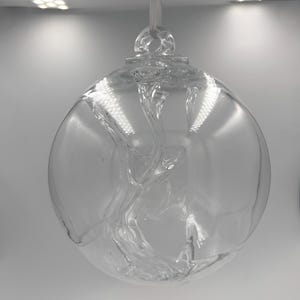 Puede incluir: Un adorno de cristal esférico y transparente que presenta un patrón interno de remolinos. El orbe hueco tiene un pequeño lazo en la parte superior para colgarlo, con una superficie lisa y transparente que refleja la luz.