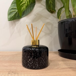 Könnte beinhalten: Ein schwarzer, gesprenkelter Diffusor für ätherische Öle mit einem goldfarbenen Kragen und Holzstäbchen. Die runde Keramikflasche steht auf einer Holzoberfläche, mit grünen Pflanzen im Hintergrund. Dieser Artikel für Raumduft ist für die Aromatherapie bestimmt.