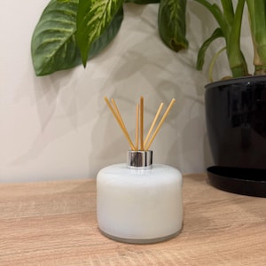 Puede incluir: Un difusor de aceites esenciales de vidrio blanco con un collar plateado y varillas de madera. El difusor está sobre una superficie de madera, con plantas verdes al fondo.