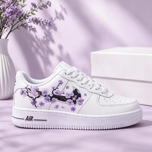 以下が含まれることがあります： 紫色の桜の枝と花で装飾された黒いスウッシュロゴが特徴の白いスニーカー。白い靴紐、側面にAIRの文字が刻印された白いソール、通気性のための穴が開いたつま先部分を備えています。