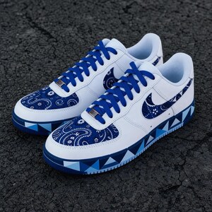 May include: A pair of white low-top sneakers featuring blue paisley patterned overlays on the toe box and side swoosh. The shoes include bright blue laces, a metallic lace dubrae, and a midsole decorated with a geometric blue triangle pattern.