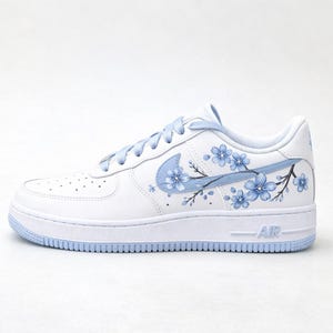 Pode incluir: Um t&ecirc;nis branco com cadar&ccedil;os azul-claros e sola azul-clara. A lateral do cal&ccedil;ado exibe um logotipo Nike azul-claro integrado a um design pintado &agrave; m&atilde;o de flores de cerejeira azul-claras em galhos marrom-escuros.