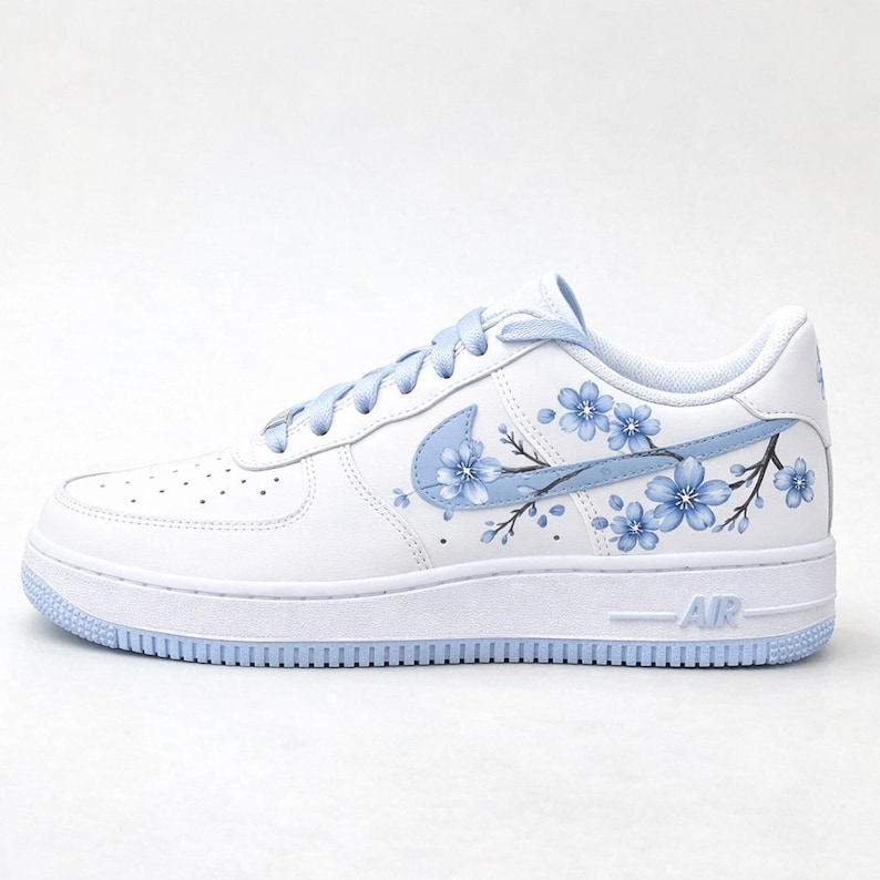 Pode incluir: Um t&ecirc;nis branco com cadar&ccedil;os azul-claros e sola azul-clara. A lateral do cal&ccedil;ado exibe um logotipo Nike azul-claro integrado a um design pintado &agrave; m&atilde;o de flores de cerejeira azul-claras em galhos marrom-escuros.