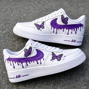 Peut inclure: Une paire de baskets blanches avec un logo swoosh violet à effet de peinture dégoulinante. Les chaussures sont décorées d'illustrations détaillées de papillons violets sur les côtés et portent le mot AIR sur la semelle et le talon.