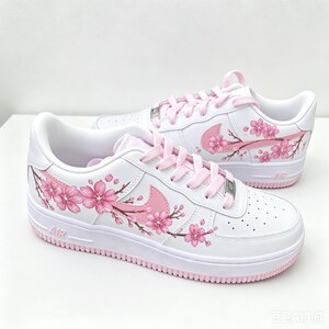 Pode incluir: Um par de t&eacute;nis brancos com atacadores rosa claro e um design de flores de cerejeira pintado &agrave; m&atilde;o em tons de rosa e castanho nas laterais. Os sapatos t&ecirc;m uma sola branca com rasto rosa, um log&oacute;tipo Nike rosa e placas prateadas.