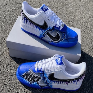 May include: A pair of white and blue Nike Air Force 1 sneakers featuring custom hand-painted designs. The shoes display black swooshes with purple and blue paint drip effects, cracked concrete textures, and the word Nike in bold black lettering.