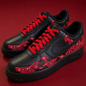 May include: A pair of black low-top sneakers featuring bright red laces and a red paisley bandana-style flame pattern along the sides and toe box. The shoes have a black leather upper, a black midsole, and a black outsole with the text AIR visible.