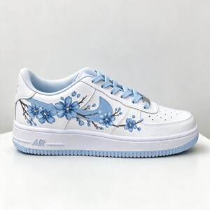 Pode incluir: Um t&ecirc;nis de cano baixo branco com um design pintado &agrave; m&atilde;o de ramos de cerejeira azuis no painel lateral. O cal&ccedil;ado inclui cadar&ccedil;os azul-claros, um swoosh da Nike azul-claro correspondente e uma entressola branca com o texto AIR em relevo.
