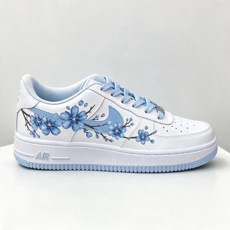 Pode incluir: Um t&ecirc;nis de cano baixo branco com um design pintado &agrave; m&atilde;o de ramos de cerejeira azuis no painel lateral. O cal&ccedil;ado inclui cadar&ccedil;os azul-claros, um swoosh da Nike azul-claro correspondente e uma entressola branca com o texto AIR em relevo.