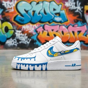 May include: White sneaker with blue and yellow graffiti-style accents. The shoe features a blue dripping paint design along the sole and a blue and yellow swoosh. The word "AIR" is printed in blue on the side.