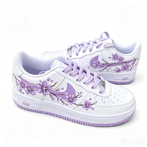 Pode incluir: Um par de t&eacute;nis brancos com um design floral de flores de cerejeira roxas pintado nos pain&eacute;is laterais e no swoosh da Nike. Os sapatos incluem atacadores roxos a condizer, uma entressola branca com o texto AIR e uma sola de borracha roxa.