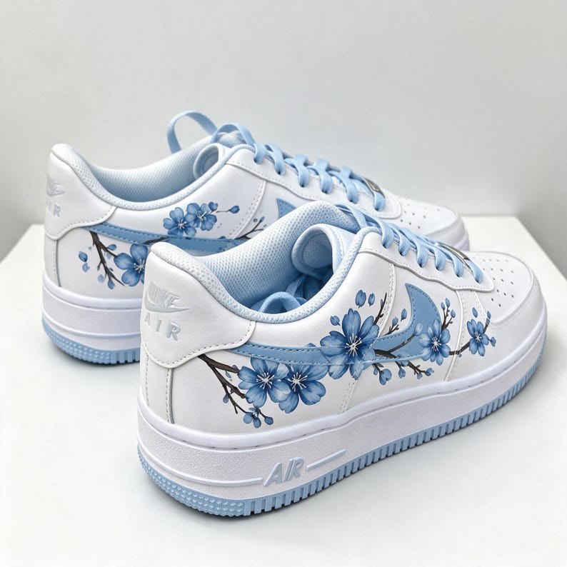 Pode incluir: Um par de t&eacute;nis brancos com ramos de cerejeira azuis pintados &agrave; m&atilde;o que se estendem pelos pain&eacute;is laterais e sobre os log&oacute;tipos Nike swoosh azul-claros. Os sapatos incluem atacadores azul-claros a condizer, solas brancas e a palavra AIR gravada no calcanhar e na sola interm&eacute;dia.
