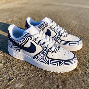 Peut inclure: Baskets blanches et bleu marine avec un motif géométrique unique. Les chaussures présentent une base blanche avec des accents bleu marine sur le swoosh, le talon et les bordures. Le design comprend des formes et des lignes complexes, créant un look élégant et moderne.