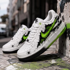 May include: A pair of white low-top sneakers featuring a black and neon green spider-themed graphic design. The shoes display a cracked pattern, black spider illustrations, and a bright green Nike swoosh logo with a matching neon green sole.