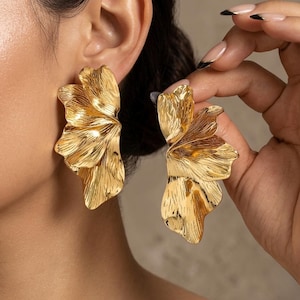 May include: A pair of large, gold-colored earrings in a floral design. The earrings have a textured surface and are shaped like stylized flower petals. The earrings are being held up to the ear.