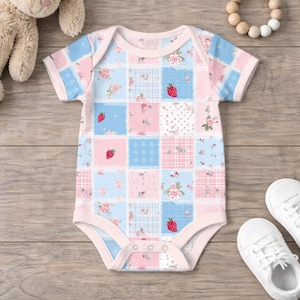 May include: A short-sleeved baby bodysuit featuring a patchwork pattern of light blue and soft pink squares. The design includes small red strawberry illustrations, floral motifs, and plaid textures, finished with light pink trim and snaps.