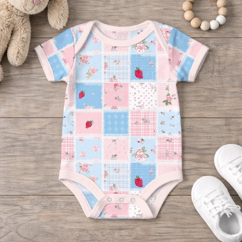 May include: A short-sleeved baby bodysuit featuring a patchwork pattern of light blue and soft pink squares. The design includes small red strawberry illustrations, floral motifs, and plaid textures, finished with light pink trim and snaps.