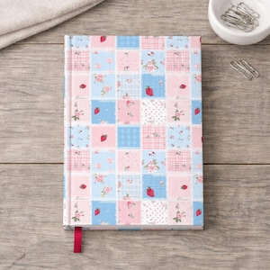 May include: A hardback notebook featuring a patchwork-style grid pattern of light blue and soft pink squares. The design includes small red strawberry illustrations, delicate floral motifs, and subtle plaid textures. A solid red ribbon bookmark extends from the bottom.