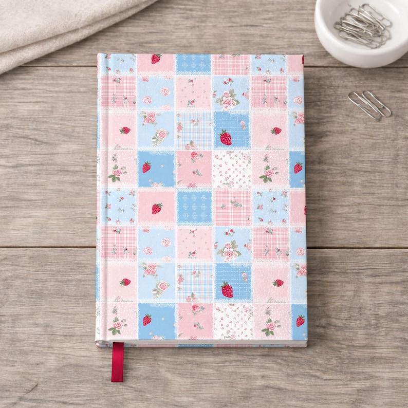 May include: A hardback notebook featuring a patchwork-style grid pattern of light blue and soft pink squares. The design includes small red strawberry illustrations, delicate floral motifs, and subtle plaid textures. A solid red ribbon bookmark extends from the bottom.