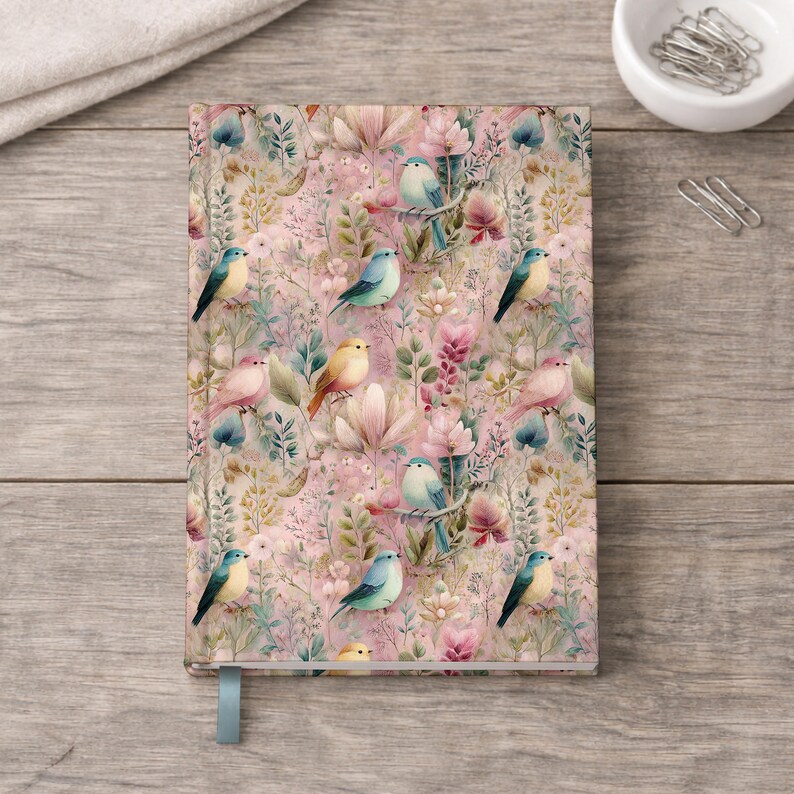 May include: A hardcover journal with a floral and bird pattern. The cover features a pink background with watercolor illustrations of birds, flowers, and foliage in shades of blue, yellow, and pink. A light blue ribbon bookmark is visible.