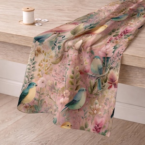 May include: A pink floral fabric with watercolor-style birds and flowers. The fabric features blue, yellow, and pink birds perched among various flowers and greenery. The fabric is draped over a wooden surface.