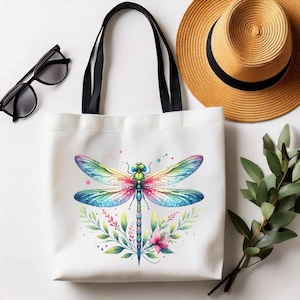 May include: White tote bag featuring a colorful dragonfly design with rainbow wings, a green body, and a floral and leaf border. The bag has black handles. Sunglasses and a straw hat are in the background.