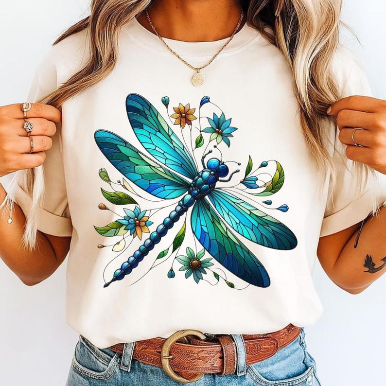 May include: Cream-colored t-shirt featuring a detailed illustration of a dragonfly with blue and green stained-glass style wings. The design includes blue and yellow flowers and green leaves, creating a nature-inspired graphic.
