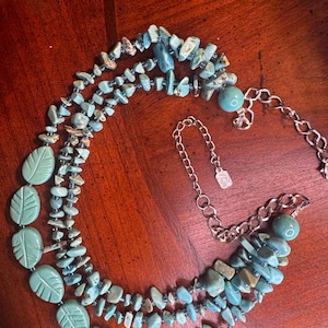 May include: A multi-strand necklace featuring turquoise-colored stone chips and leaf-shaped beads. The necklace has a silver-toned chain and clasp. The stones are a mix of light and dark blue, with some brown accents. The necklace is designed for a layered look.