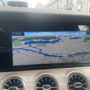 Puede incluir: Panel de instrumentos de un coche con una pantalla digital que muestra un mapa de Riga, Letonia. La pantalla muestra la hora 12:03. El salpicadero tiene un acabado de veta de madera y rejillas de ventilación circulares.