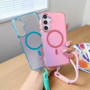 Puede incluir: Dos fundas transparentes para teléfono, una azul y otra rosa, con un diseño circular y un símbolo de carga magnética. La funda rosa tiene un cordón rosa y la funda azul tiene un cordón turquesa. Las fundas están sujetas por una mano.