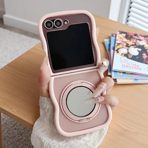 Puede incluir: Una funda de teléfono rosa con espejo y lente de cámara integrados. La funda está abierta, revelando una pantalla negra. El diseño tiene una forma suave y redondeada. La funda del teléfono está sobre una superficie de madera.