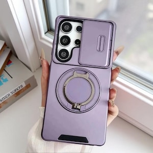 May include: A lilac phone case featuring a camera cover and a ring holder. The case has a glossy finish and a rectangular shape. The ring holder is silver and circular. The camera area has three lenses.